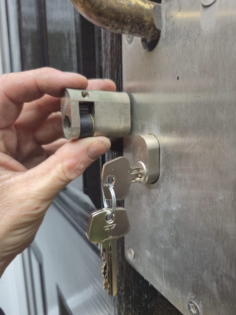 An oval half-cylinder held against the brushed-steel plate of a communal entrance door, keys in the lock