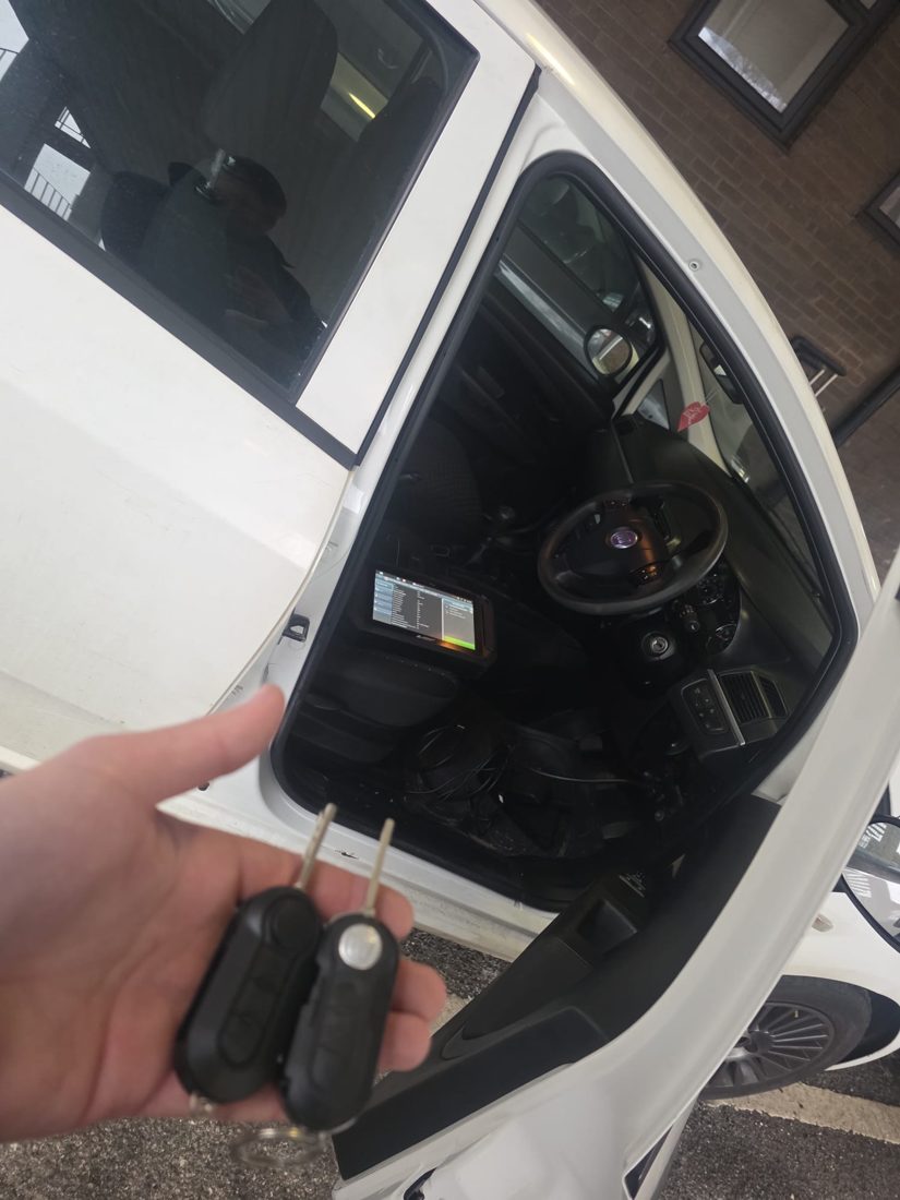 A newly cut and programmed car key held at an open car door, with diagnostic equipment plugged in on the seat