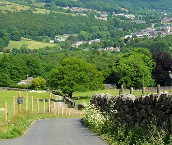 A country lane dropping between drystone walls into the Calder Valley near Halifax, with a hillside township spread across the far side