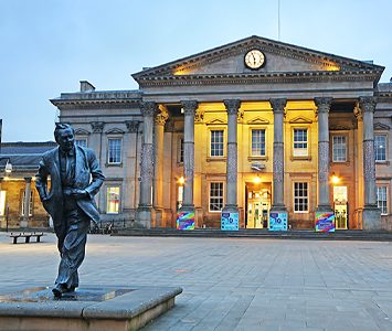 The grand classical frontage of Huddersfield railway station at dusk, with the statue of Huddersfield-born Prime Minister Harold Wilson on St George's Square