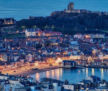 Scarborough's South Bay lit up at dusk, with the ruins of Scarborough Castle on the headland above the old town
